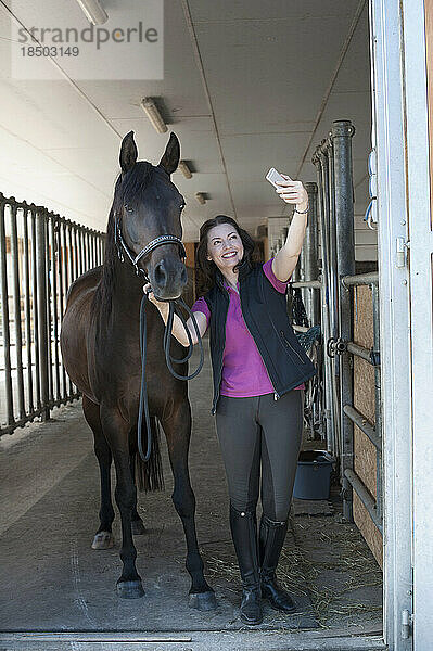 Reife Frau macht ein Selfie mit ihrem braunen Pferd im Stall und lächelt  Bayern  Deutschland