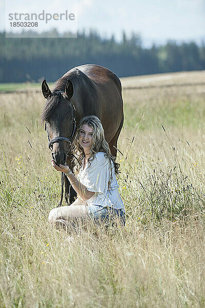 Junge Frau streichelt ihr braunes Pferd  sitzt auf der Wiese und lächelt  Bayern  Deutschland