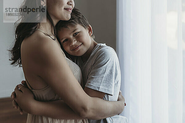 School-aged son being embraced by mother in natural light studio