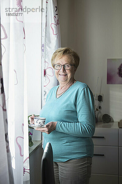Lächelnde alte Frau am Fenster mit einer Tasse Kaffee