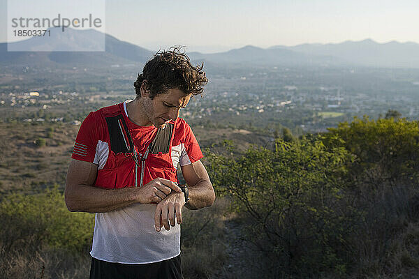 Ein sportlicher männlicher Trailrunner blickt auf seine Uhr  während die Sonne untergeht  nachdem er auf den Trails in der Nähe der Berge von El Arenal  Hidalgo  Mexiko  gelaufen ist.