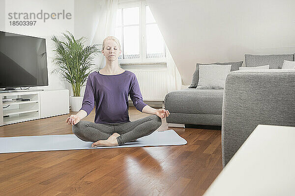 Junge Frau macht Lotus-Pose-Yoga auf einer Gymnastikmatte im Wohnzimmer  Bayern  Deutschland