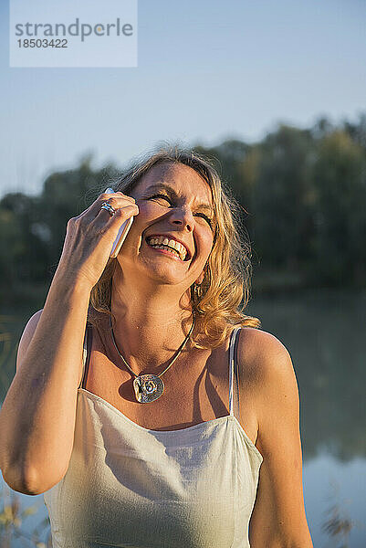 Fröhliche Frau lacht  während sie am Flussufer mit dem Smartphone spricht  Bayern  Deutschland