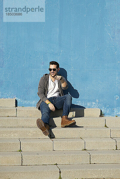 Vorderansicht eines jungen bärtigen Mannes  der auf einer Außentreppe sitzt  wegschaut und lächelt