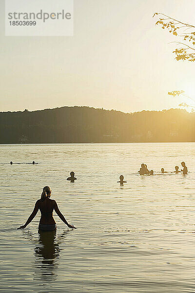 Reife Frau spaziert bei Sonnenuntergang im See und ihre Freunde schwimmen im See  Bayern  Deutschland