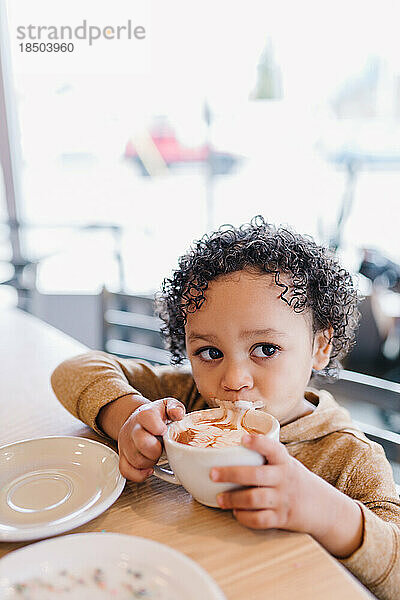 Entzückendes biraziales Kleinkind  das an einer Tasse Kakao nippt