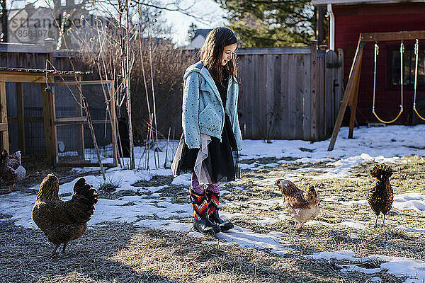 Kleines Mädchen in Kostüm und Winterstiefeln blickt auf Hühner herab