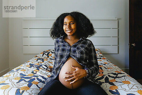 Eine junge Frau sitzt auf dem Bett und hält den schwangeren Bauch