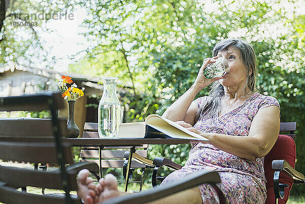 Ältere Frau trinkt Wasser  während sie im Garten ein Buch liest  Altötting  Bayern  Deutschland