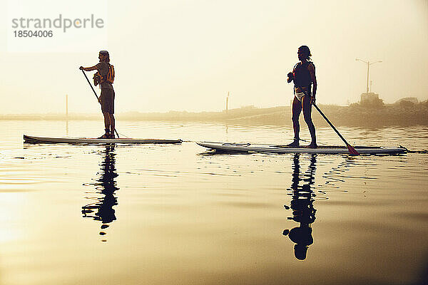 Zwei Freunde beim Standup-Paddle-Boarding an einem nebligen Morgen bei Sonnenaufgang