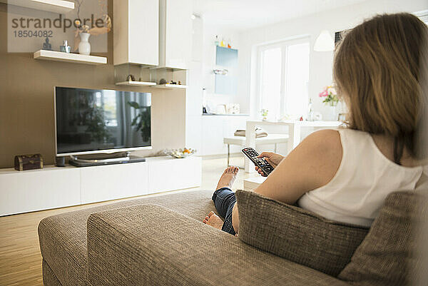 Schwangere Frau sitzt auf dem Sofa und schaut mit Fernbedienung fern  München  Bayern  Deutschland