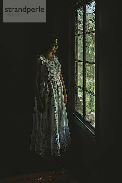 Erwachsene Frau in weißem Kleid blickt aus dem Fenster in natürlichem Licht