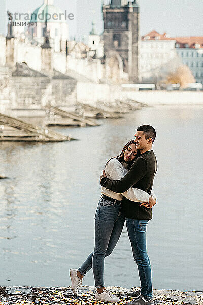 Mann und Frau umarmen sich in der Nähe von Fluss und Brücke im Zentrum von Prag