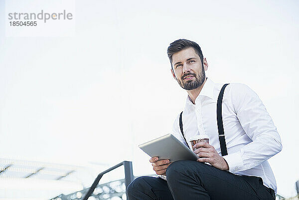 Geschäftsmann sitzt auf der Treppe und arbeitet mit Kaffee am digitalen Tablet  München  Bayern  Deutschland