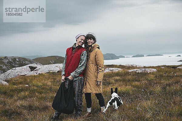 Queeres Paar und Hund stehen im schottischen Hochland mit Meerblick