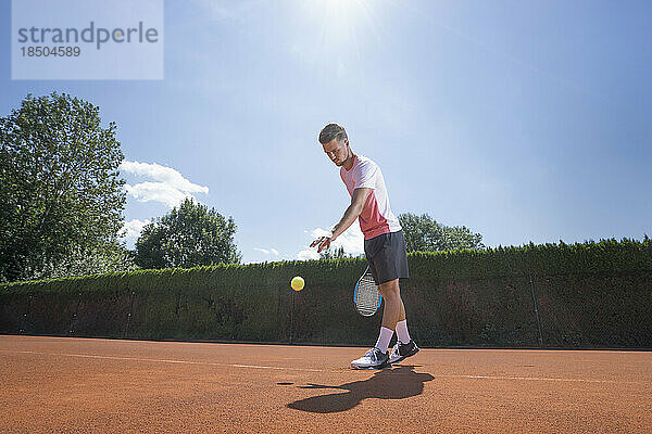 Junger männlicher Tennisspieler bereitet sich darauf vor  den Ball auf dem sonnigen roten Tennisplatz in Bayern  Deutschland  zu servieren