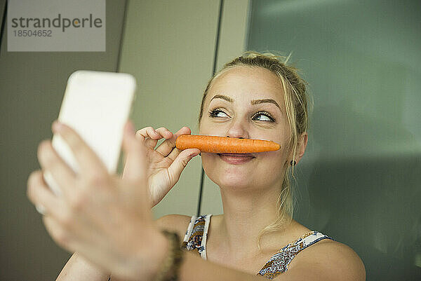 Junge Frau macht ein Selfie und macht einen Schnurrbart mit einer Karotte im Gesicht  München  Bayern  Deutschland
