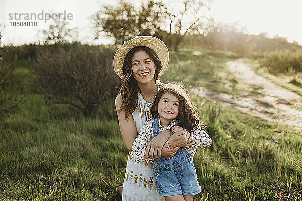 Porträt einer glücklichen jungen Mutter und Tochter  die auf einer Wiese im Gegenlicht lächelt