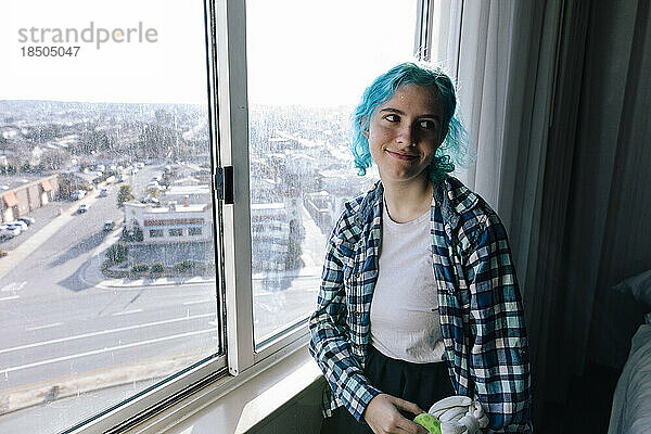 Teenager-Mädchen mit blauen Haaren lächelt neben schmutzigem Hotelfenster