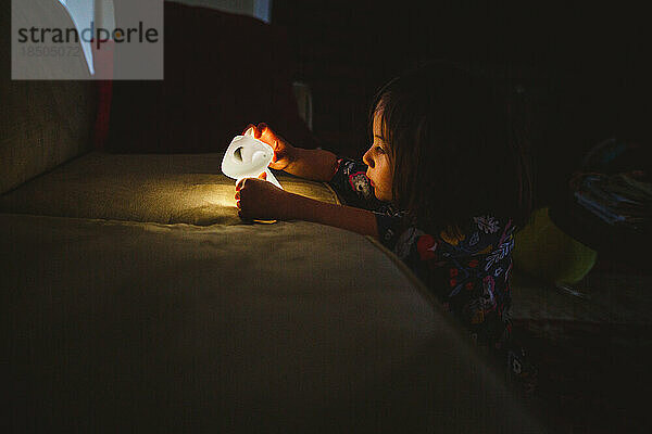 A little girl sits in the dark with her face lit up by a small lamp