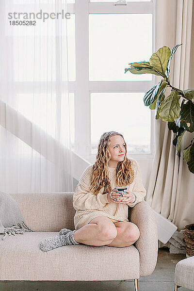 Nachdenkliche junge Frau im kuscheligen Pullover auf der Couch und blickt aus dem Fenster