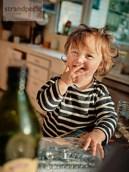 Kleinkind mit lockigem Haar bedeckt den Mund mit der Hand  während es in der Küche lacht