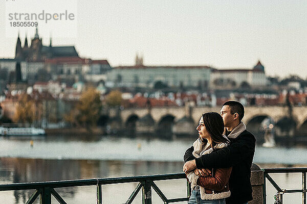 Mann und Frau umarmen sich in der Nähe von Fluss und Brücke im Zentrum von Prag