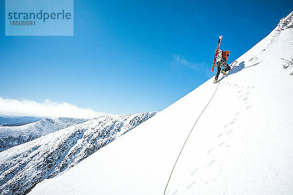 Mann geht mit Eiskletterwerkzeug und Skiern auf dem Rücken bergauf