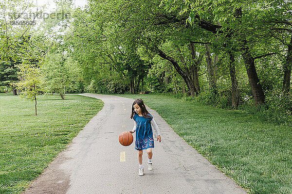 A little girl dribbles basketball alone down long wooded pathway