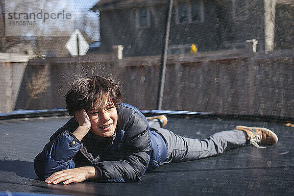 A smiling boy lays on a trampoline outside in winter