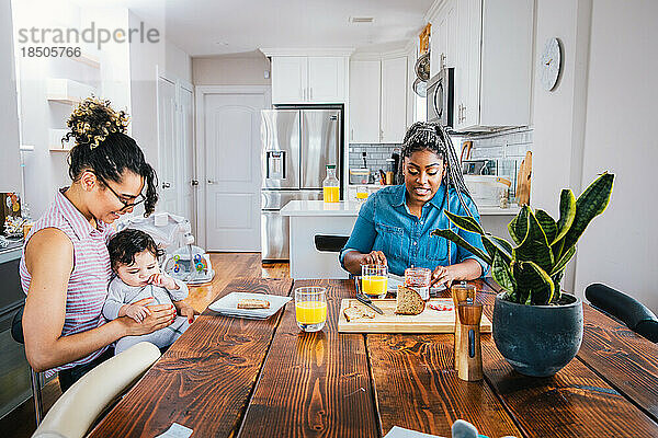 Glückliche Mutter spielt mit ihrem kleinen Jungen  während ihre Freundin am Tisch das Frühstück zubereitet