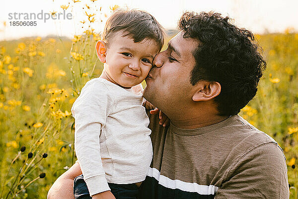 Kleiner Sohn lächelt  während Latino-Vater draußen auf dem Feld einen Kuss auf die Wange gibt