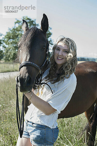 Junge Frau streichelt ihr braunes Pferd  steht auf der Wiese und lächelt  Bayern  Deutschland