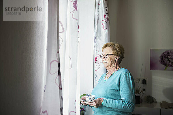 Lächelnde alte Frau  die mit einer Tasse Kaffee aus dem Fenster schaut