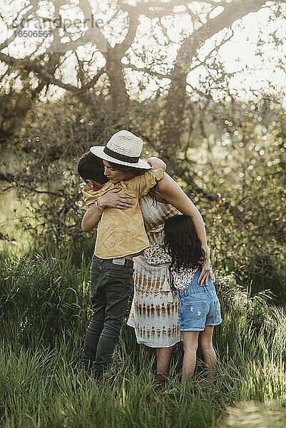 Ganzkörperansicht einer Mutter  die Sohn und Tochter in einem von hinten beleuchteten Feld umarmt