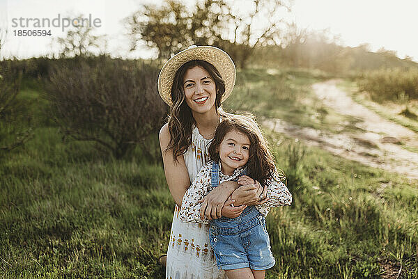 Porträt einer glücklichen jungen Mutter und Tochter  die auf einer Wiese im Gegenlicht lächelt
