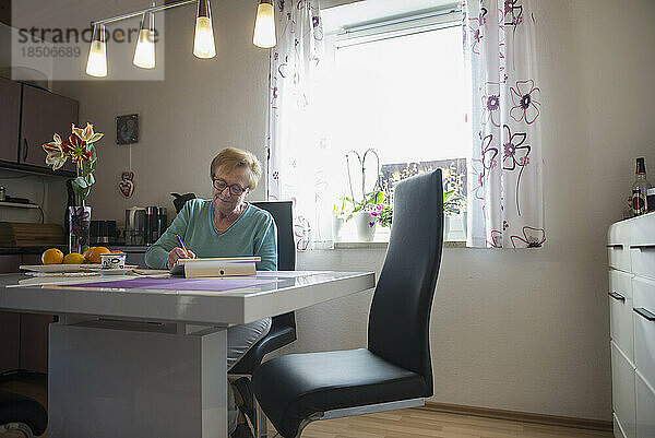 Alte Frau schaut am Esstisch auf ein digitales Tablet und schreibt in ein Buch