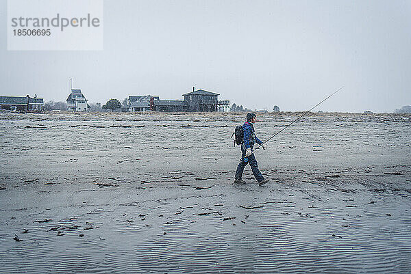 Frau läuft mit Fliegenfischerausrüstung im Schnee über den Strand