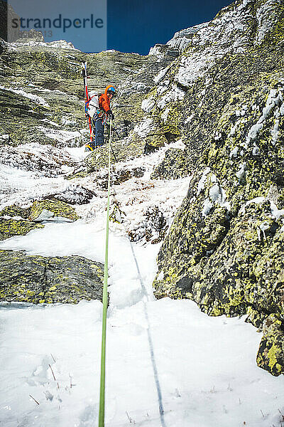 Mann Mixed-Eiskletterer mit Skiern auf dem Rücken beim Sichern auf Felsen