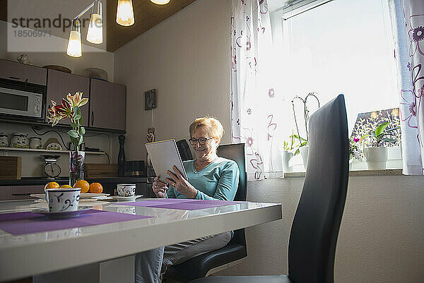 Glückliche alte Frau schaut am Frühstückstisch auf ein digitales Tablet