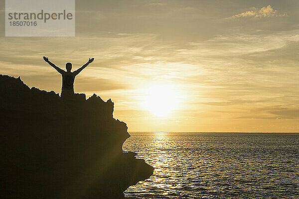 Silhouette eines Mannes  der bei Sonnenuntergang mit ausgestreckten Armen auf einer Klippe steht  Nusa Lembongan  Bali  Indonesien