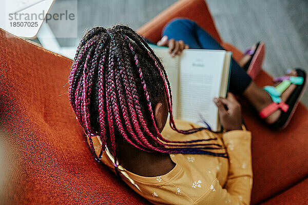 Junges afroamerikanisches Mädchen liest ein Buch auf einem orangefarbenen Stuhl
