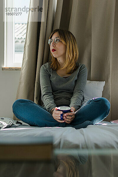 Junge Frau mit einer Tasse Kaffee in ihrem Schlafzimmer.