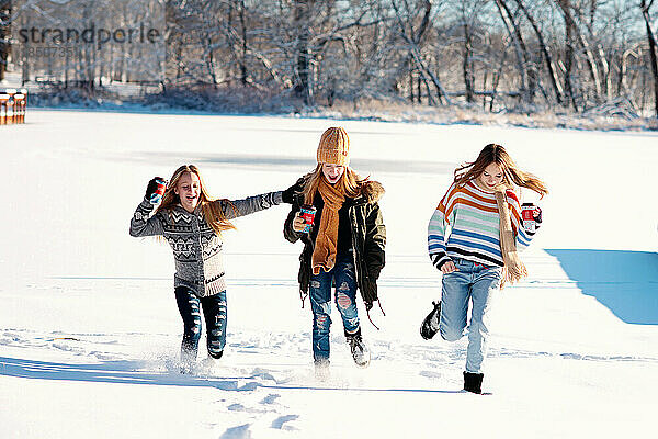 Drei Teenager-Mädchen laufen an einem Wintertag im Schnee auf einem zugefrorenen See.