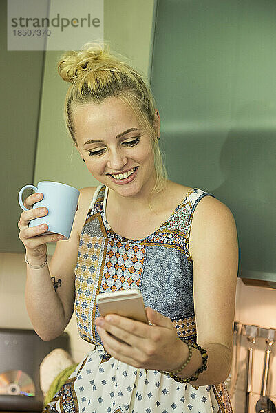 Schöne junge Frau schaut auf ihr Smartphone und trinkt Kaffee in der Küche  München  Bayern  Deutschland