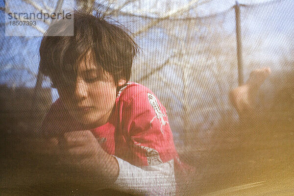 Blick auf einen Jungen  der durch ein Trampolinnetz im Sonnenlicht liegt