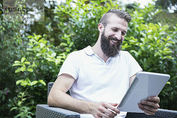 Glücklicher junger Mann sitzt auf einem Stuhl und nutzt ein digitales Tablet im Garten  Bayern  Deutschland