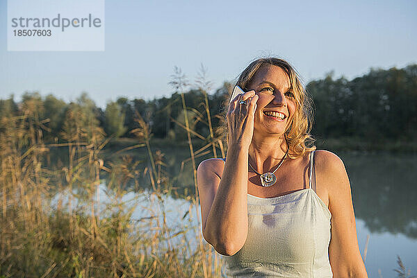 Fröhliche Frau lächelt  während sie am Flussufer mit dem Smartphone spricht  Bayern  Deutschland