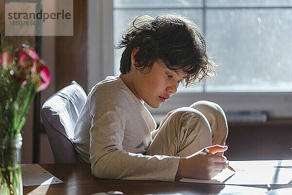 Ein Junge mit zerzausten Haaren sitzt am Tisch und schreibt mit Bleistift auf Papier