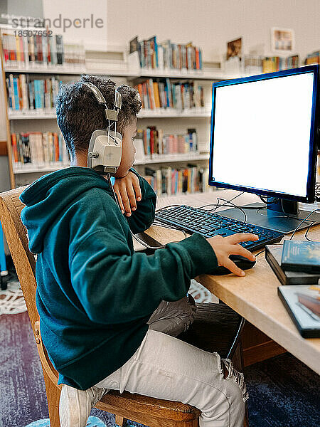 Kleiner Junge mit Kopfhörern benutzt Computer in öffentlicher Bibliothek